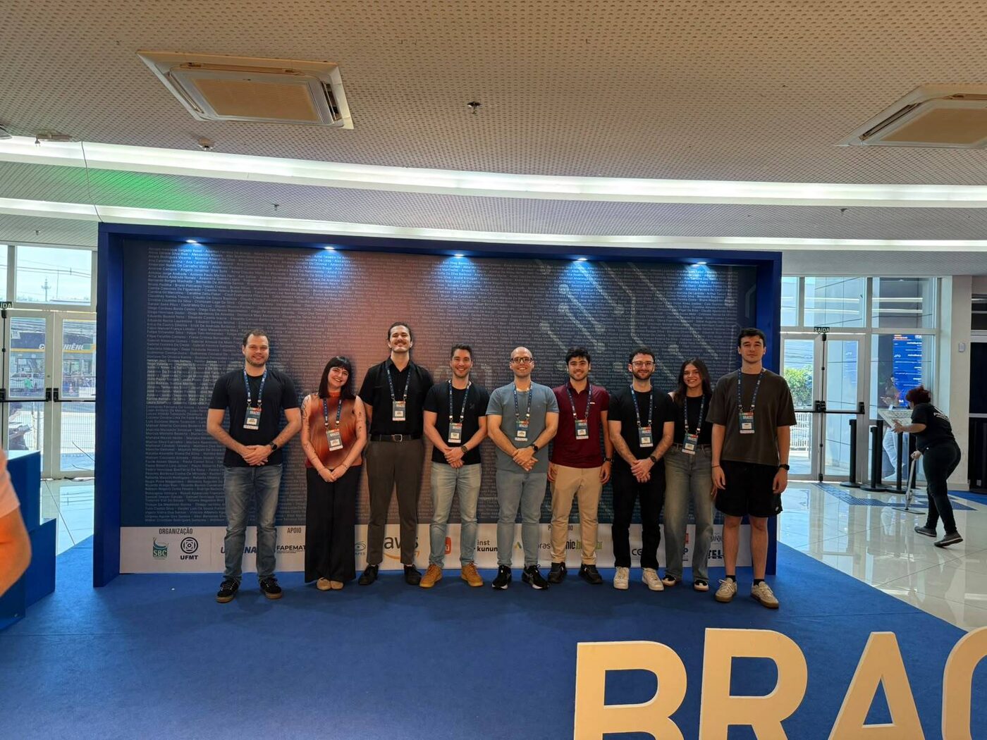 Group photo of nine MALTA members in front of the BRACIS 2026 wall of participant names